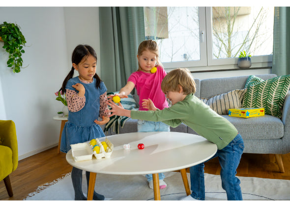 Drie kinderen spelen het Haba Spellen Eierdans behendigheidsspel met speelgoed eieren en lepels rond een witte tafel in een lichte woonkamer, terwijl op de achtergrond een bank, planten en een bordspel te zien zijn.