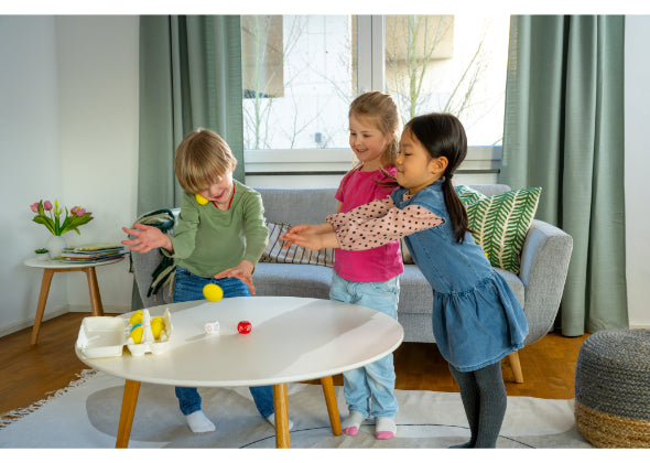 Drie kinderen spelen Eierdans van Haba Spellen en gooien balletjes op een witte tafel. Lachend en plezier makend genieten ze van dit behendigheidsspel in een woonkamer met een grijze bank, groene gordijnen en bloemen op de achtergrond.
