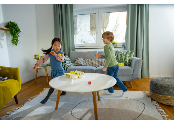 Twee jonge kinderen spelen energiek in een lichte woonkamer met een ronde witte tafel, speelgoedkuikens en een rode dobbelsteen - perfect om Eierdans van Haba Spellen te spelen. De ruimte is voorzien van groene gordijnen, een grijze bank en speelse decoraties.
