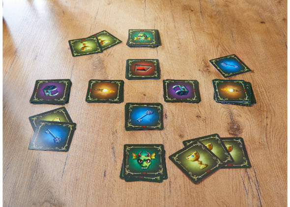 Verschillende kleurrijke speelkaarten van Dragonella van Haba Spellen, een vrolijk kinderbordspel met sleutels, bekers, monsters en pijlen, liggen verspreid in groepjes en stapeltjes op een houten tafel.