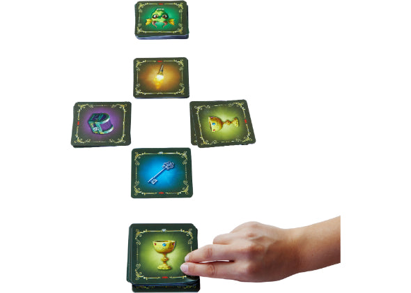 Een hand plaatst een kaart met een gouden bokaal uit Haba Spellen's Dragonella tussen vijf geïllustreerde kaarten met verschillende voorwerpen, allemaal gerangschikt op een wit vlak.